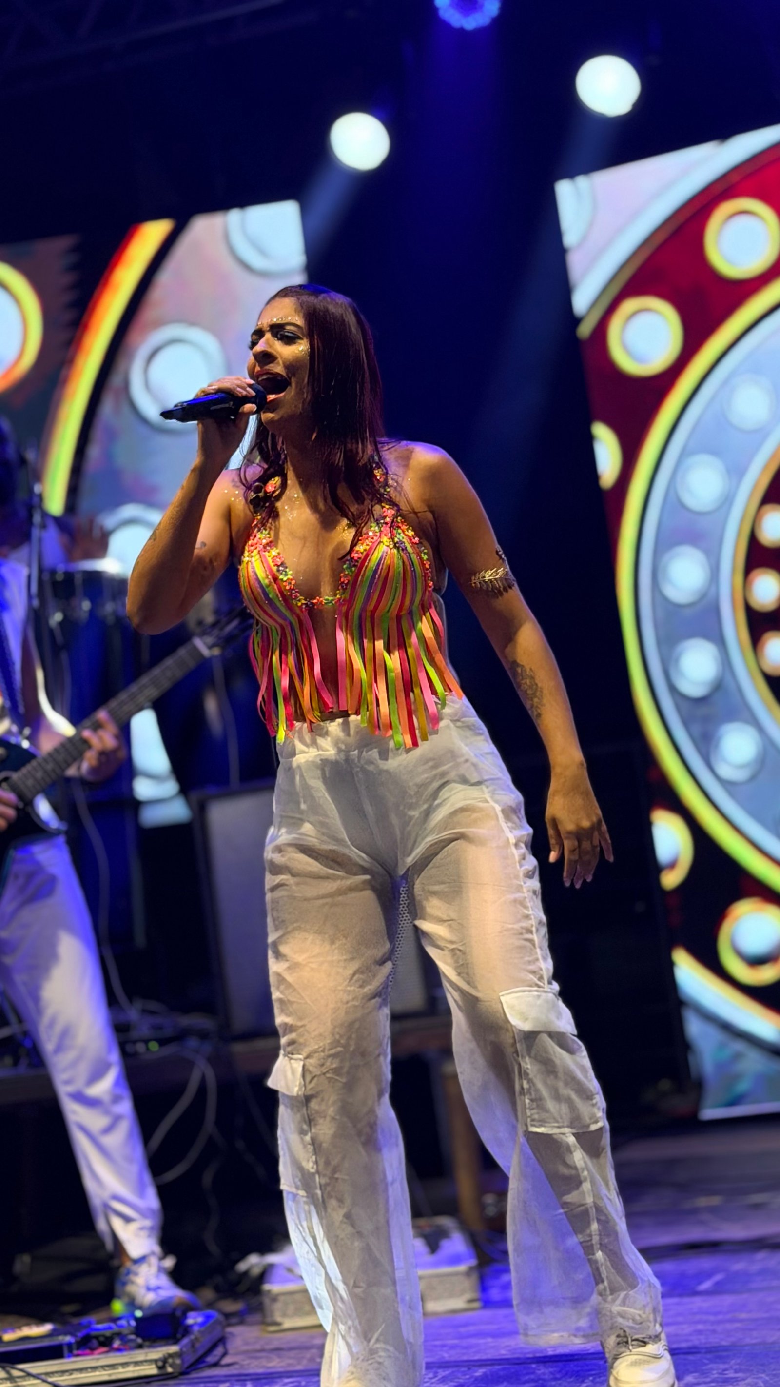 Carol Ferreira cantando ao vivo com roupa colorida e cenário iluminado